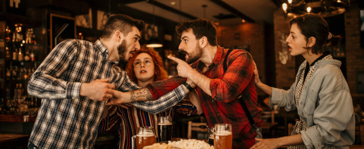 A two young adult bearded guys having an argument, getting a bit physical while in the pub with their girlfriends, that are trying to separate them. Both are wearing a plaid shirts, treating each other