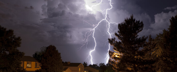 A massive thunderhead pours rain and lightning over suburban Denver homes, Colorado. Trees are silhouetted and homes are lit by incandescent light. The thunderhead is lit by huge lightning bolts reaching from the top of the clouds to the ground.