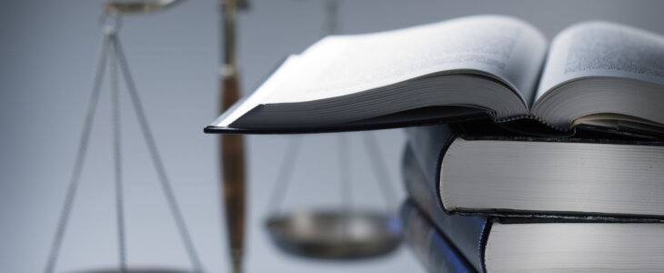 A stack of law books stands in front of a justice scale that is slightly out of focus. On top of the stack is an open law book.